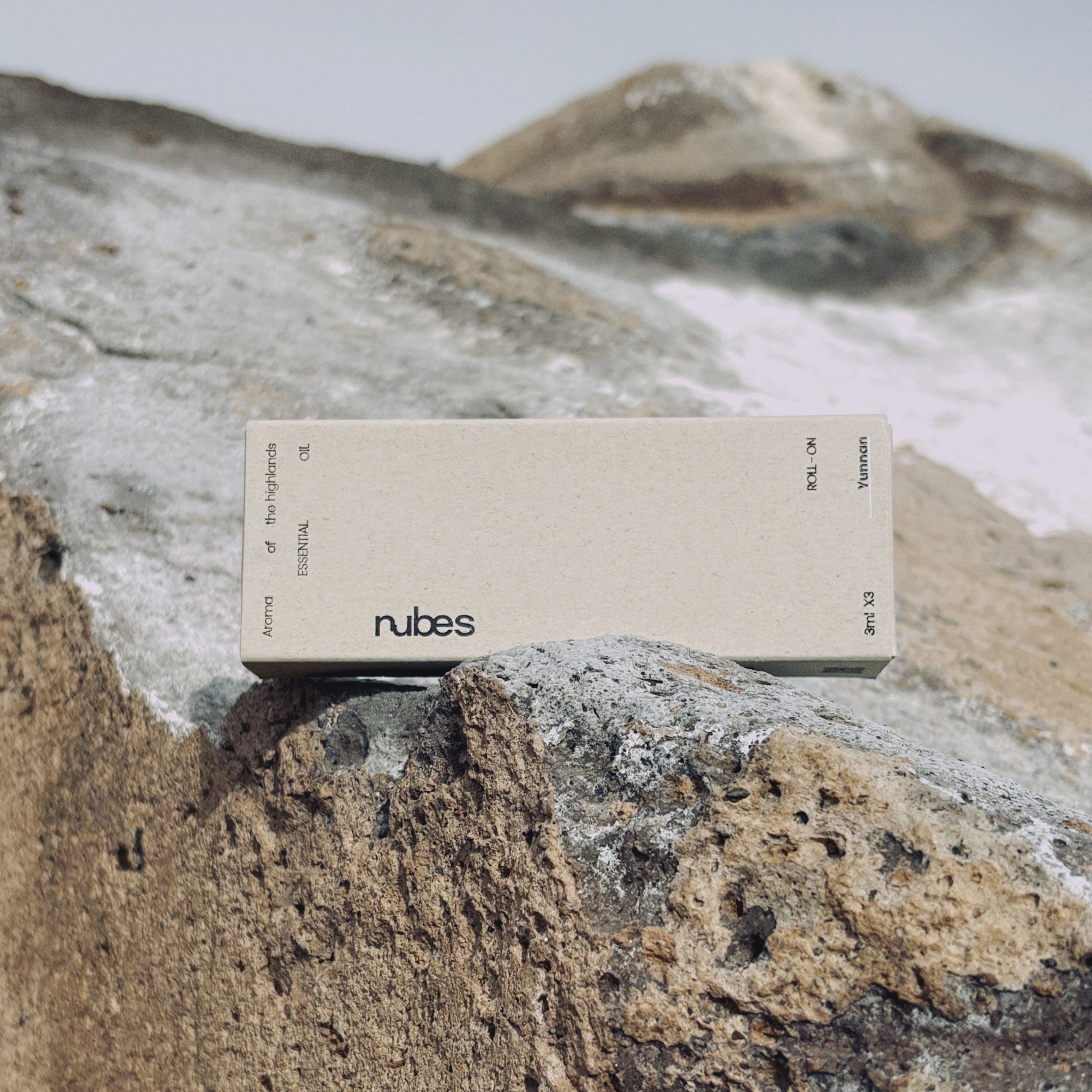 A Nubes Essential Oil Roll-On box placed on rugged rocks against a mountainous backdrop, emphasizing its natural and earthy essence.