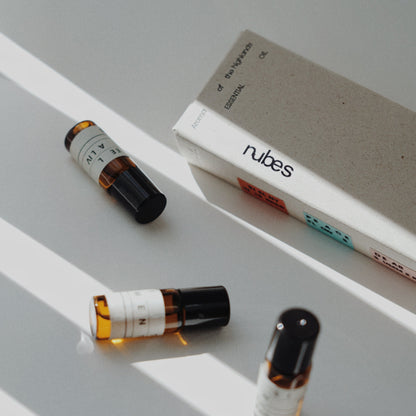 Three Nubes roll-on essential oil bottles next to eco-friendly packaging on a white surface with soft diagonal shadows.